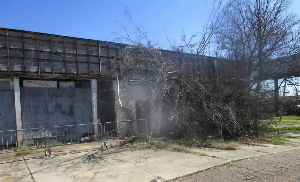 Boarded-up front of the vacant commercial building, with brush piled along the fence, before demolition
