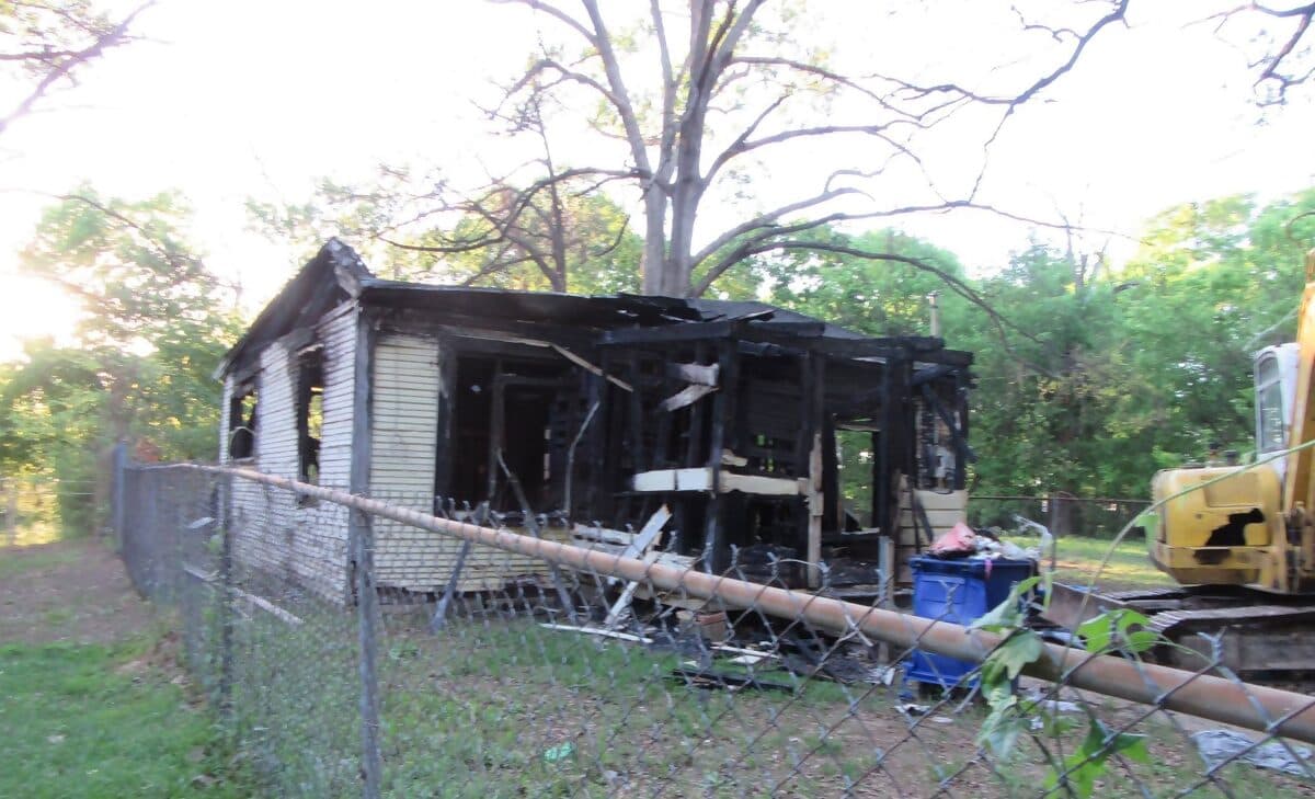 Burned house behind a chain-link fence, with the excavator moving in